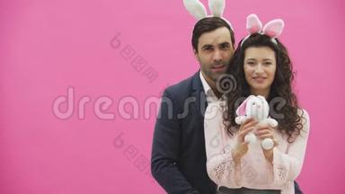 粉红色背景的年轻创意夫妇。 头上戴着黑黑的耳朵。 在这期间，一个妻子抱着一只软玩具兔子，一只